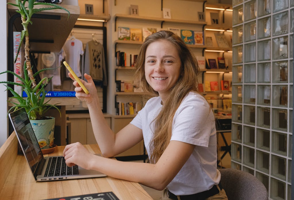 A smiling young woman uses her laptop and smartphone in a cozy cafe setting, creating a warm and inviting atmosphere.